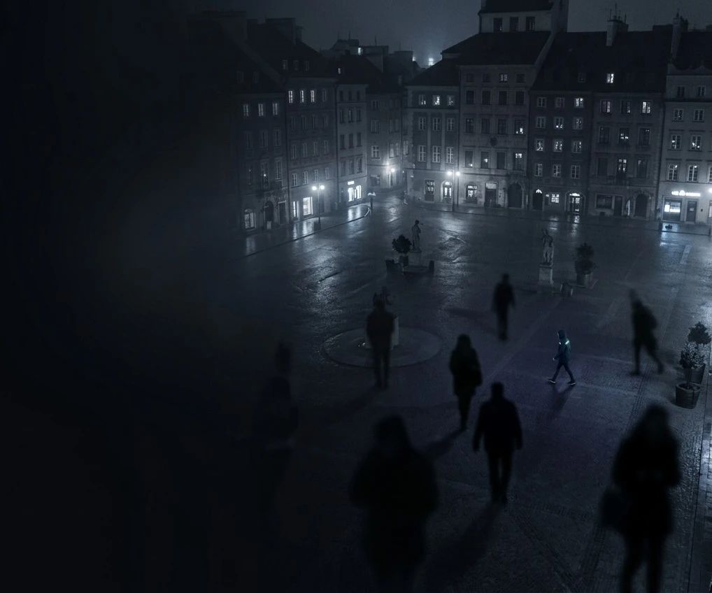 Plaza de una ciudad de noche con personas caminando en penumbra y una sola persona iluminada que destaca entre todas